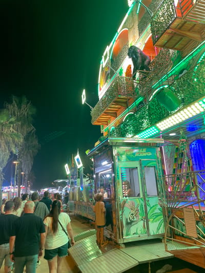 An outdoor nighttime scene captures a crowded amusement park walkway with a colorful, lit attraction in the background, identified by its "JUNGLE KING" signage. The attraction features a large gorilla statue and jungle-themed decorations, with bright green and yellow lights illuminating the structure. A queue of people, appearing to be of various ages and ethnicities, walks along the pathway. Some individuals are looking at the attraction, while others are walking forward. The ground is paved, and palm trees are visible on the left side of the image, suggesting a warm climate. A sign reading "Normas" (Spanish for "Rules") is attached to the side of the attraction. The overall atmosphere suggests a lively evening at a fair or amusement park in Canet de Mar, Spain.