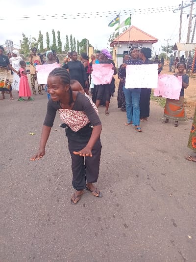 A group of women are standing on a paved road in front of a building in Ungwan Tanko, Nigeria. They are holding signs and appear to be protesting. The woman in the foreground is wearing a black shirt and black pants and has a child on her back. She is facing the camera and holding out her hands. The women in the background are wearing various clothes and holding signs. The building behind them has a green roof and a sign in front of it. There are trees and power lines in the background. The scene appears to be a protest, possibly about a local issue.