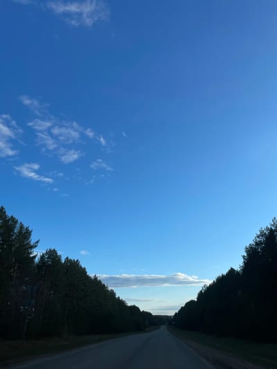 The image shows a road surrounded by trees under a blue sky with scattered clouds. The location is Средний Постол, Russia.