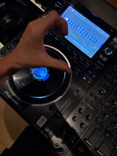 This media file is a close-up, overhead shot of a Pioneer DJ CDJ-3000 multi-player. The device is primarily black with illuminated blue and white elements on its screen and buttons. A person's hand, adorned with rings, is visible in the foreground, interacting with the DJ equipment. The hand is positioned as if to manipulate the jog wheel or touch the screen.

The screen displays a waveform, track information including the title "Jaaron (Original Mix)", track length, BPM (123.0), and a progress bar indicating 03:55 remaining. Other visible text on the DJ unit includes "Pioneer DJ", "CDJ-3000", "MASTER PLAYER", "TEMPO", "FILTER", "BEAT SYNC", "VINYL", and various button labels like "LOOP", "KEY SHIFT", "BEAT JUMP", "HOT CUE", "SEARCH", and "PLAYLIST". There is also a QR code sticker on the unit with text in what appears to be multiple languages.

The scene is dimly lit, suggesting it might be indoors or nighttime. The focus is entirely on the DJ equipment and the hand interacting with it, implying the activity is DJing or preparing to DJ. No specific location cues are evident beyond the make and model of the equipment. The overall impression is one of focused activity and the technical aspect of music performance.
