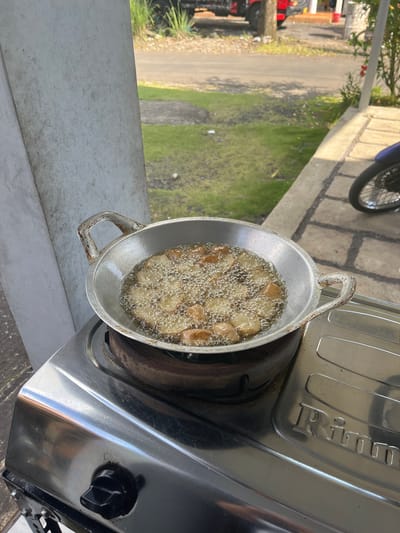 In Jambearum, Indonesia, a close-up shot shows food being fried in a wok on a gas stove. The wok, made of aluminum, sits atop a rusty burner. The food, appearing to be small, golden-brown pieces, is submerged in bubbling oil, creating a lively surface. The gas stove is stainless steel, with a black knob visible on the front. In the background, a paved area with grass and a road is visible, with a red truck and some trees suggesting an outdoor setting.