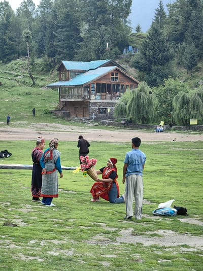 A group of people are gathered in a grassy field in front of a wooden building with a blue roof in Solang, India. In the foreground, a woman wearing a red and gold traditional outfit is kneeling and presenting a basket of pink flowers to another woman dressed in a blue and gray checkered garment. Beside them stands a man in a dark traditional outfit. To the right, a man in a light blue shirt and beige pants faces away from the camera. In the background, several other individuals are scattered throughout the field and near the building. The environment appears to be mountainous, with trees and a hillside visible behind the structure. Bags and other items are present on the ground in the foreground.
