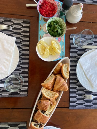 The image shows a table setting with a variety of food items. In the center of the table is a white oval dish filled with croissants and a few pastry-like items. To the right of this dish is a white napkin, a glass, a silver fork, and a white plate. To the left is a black and white placemat, a white napkin, a white plate, a glass, and another black and white placemat. On the table is a blue and white ceramic serving dish filled with parsley and a few donuts. The dish is also decorated with a white ceramic rooster, a white ceramic container, a glass bottle, and a bowl filled with red, diced tomatoes. The scene is shot from a bird's-eye view. The coordinates provided correspond to a location in Rye, New York, but the image itself does not provide any evidence of the location.