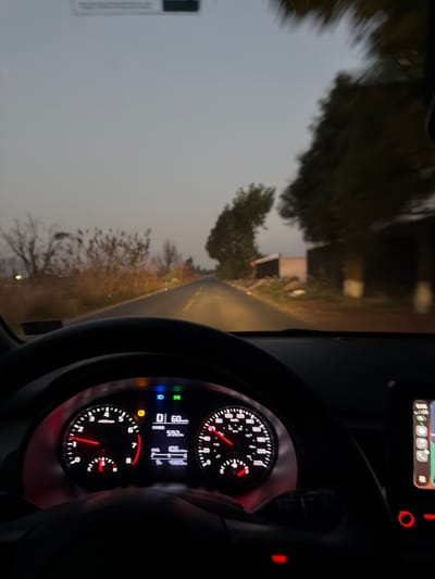 The image is taken from inside a moving vehicle, likely a car, showing the dashboard and the road ahead. The speedometer indicates a speed of approximately 60 km/h. The road is paved and appears to be winding through a somewhat rural or suburban area, with trees and a building visible on the side. The lighting suggests it is either early morning, late evening, or a cloudy day. Given the camera angle, the driver is the subject, and the action is driving. Based on the details available in the photo and with the details of the question, determining the specific location in San Pedro Cholula, Mexico, is impossible without additional information or clearer landmarks in the background.