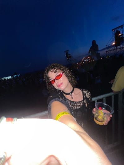 An outdoor, nighttime image captures a young woman with curly brown hair and red-tinted sunglasses. She is wearing a black, fishnet-style top and a choker necklace with a cross pendant. A yellow wristband is visible on her left arm, reading "DAY 1". She holds a clear plastic cup containing a drink with a cherry and possibly a slice of fruit. The background reveals a dark blue, twilight sky with distant stage lights and structures, suggesting an outdoor concert or festival. A crowd of people is visible in the mid-ground, and a metal railing is in the foreground. The scene is consistent with an event taking place in Donji Štoj, Montenegro, known for its outdoor music festivals held near the coast during the summer.