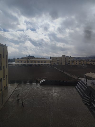 An outdoor scene in Jibb as-Safa, Syria, captured on an overcast day with a cloudy sky showing patches of brighter clouds. The image is dominated by institutional architecture and open ground.

In the foreground, a wet, dark-colored paved courtyard, appearing to be made of cobblestones or pavers, occupies the lower third of the frame. Two individuals, appearing as dark figures, walk on this pavement; one is closer to the left edge, facing right, and another is further right, facing left. To the far left, the side of a multi-story, yellow-toned building with rectangular windows is partially visible. To the right of the paved area, a series of dark steps ascends to a raised section, which features a white or light-colored awning under which one or two indistinct figures are present. A dark, stepped retaining wall or planter separates the paved area from a large, dark brown, possibly muddy or sparsely vegetated field that spans the mid-ground.

Across this field, a dirt or gravel path runs diagonally from the left-center towards the right, with several small clusters of people, appearing as dark silhouettes, walking along it. In the background, a cluster of multi-story, yellow-toned buildings with numerous windows and various architectural details, including turrets and rooftop equipment like antennas, stretches horizontally. Beyond these buildings, in the far background to the right, hazy outlines of distant hills or mountains are discernible, along with some indistinct industrial structures.