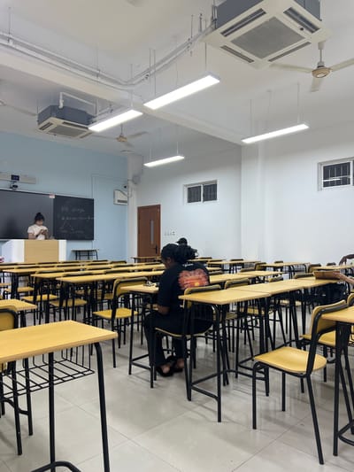 The image depicts a classroom environment, likely in a school or university setting, in Liandu District, China. The room is equipped with rows of desks and chairs, illuminated by fluorescent lighting and ceiling fans. A teacher or student stands at the front of the classroom, near a blackboard displaying some writing. Several students are seated at desks, some appearing to be engaged in learning activities. The room has a clean and organized appearance, with white walls and a tiled floor. The overall atmosphere suggests a typical educational setting.