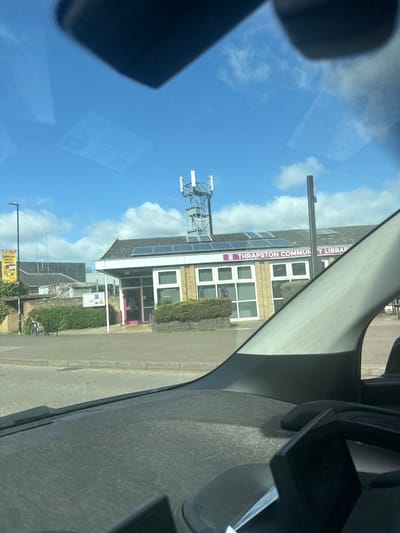 The image shows a view from inside a car, looking out towards a building identified as the "Thrapston Community Library" in Thrapston, a town within the North Northamptonshire unitary authority of Kettering, United Kingdom. The building has a brick facade with multiple windows. A communications tower is visible on the roof. A street scene is in front, with signs and vegetation visible. The sky is blue with white clouds.