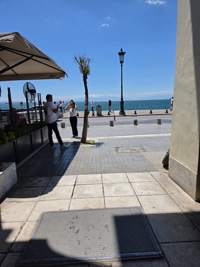 The image depicts a sunlit street scene in Thessaloniki, Greece, likely along the waterfront promenade, given the presence of the sea and distant islands or landmasses. The foreground is dominated by a patterned concrete pavement, with a metallic grate set into it, casting a strong shadow. To the left, a white outdoor umbrella of a cafe or restaurant extends into the frame, with a black industrial fan visible beneath it. A man in a white t-shirt and dark pants stands on the pavement, facing away from the camera and holding a phone. Next to him, a woman in a white t-shirt and black pants also holds a phone, seemingly taking a photograph or video.

Behind them, a narrow tree with sparse foliage stands on the sidewalk. Further into the background, the turquoise sea stretches out under a clear blue sky dotted with a few white clouds. A black lamppost with ornate detailing stands on the edge of the sidewalk, overlooking the sea. Several other people are visible in the mid-ground, some walking along the promenade and others standing near the water's edge, also appearing to be engaged with their phones. A light-colored building wall occupies the right side of the frame, casting a significant shadow across the pavement in the foreground. The overall atmosphere is one of a pleasant, leisurely day in an urban coastal environment.