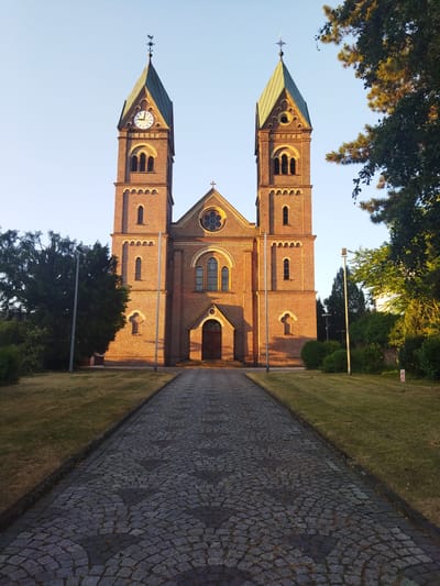 The image shows St. Andrew's Church (St. Andreas Kirche) in Leverkusen, Germany. The church is constructed of red brick and features two prominent towers with green spires. A clock is visible on the left tower. A cobblestone path leads to the main entrance of the church, with grass lawns on either side. Trees and shrubbery surround the building, and the sky is clear and blue. The sun is casting long shadows, suggesting it is either early morning or late afternoon. There are no people visible in the image.