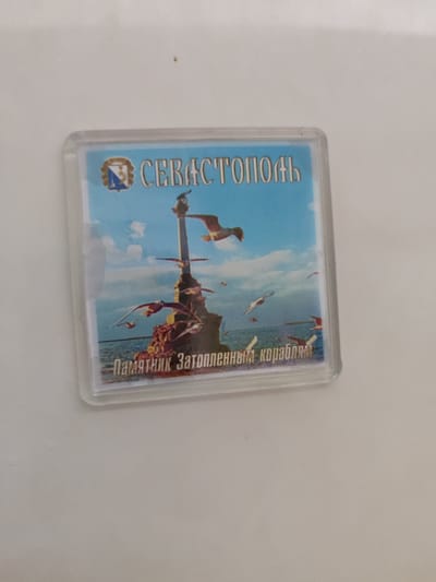 The image shows a magnet with a picture of the Monument to the Scuttled Ships in Sevastopol. The monument is depicted in the foreground, with several seagulls flying around it. The sea and the sky are visible in the background. Text on the magnet reads "СЕВАСТОПОЛЬ" and "Памятник Затопленным кораблям". This image is a souvenir and is not related to Bondarivka, Ukraine.