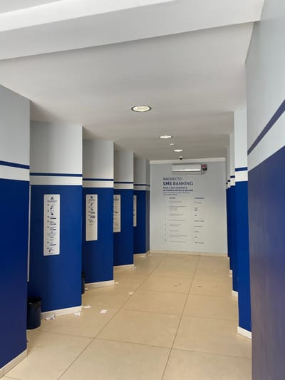 The image displays an empty, well-lit indoor corridor or service area, characteristic of a banking facility, located in Luanda, Angola. The space is dominated by a clean and modern blue and white color scheme.
The walls and ceiling are white, accented with bold horizontal and vertical royal blue stripes. The floor is tiled with light beige or cream-colored rectangular tiles, showing minimal wear.
Along the right side of the corridor, a series of individual service stations or cubicles are arranged, providing a sense of privacy. These partitions are primarily blue with white tops. Each cubicle features a white informational sign, visibly labeled "A", "B", and "C" (from the foreground to the background). These signs contain pictograms and brief text, likely instructing users on how to access or utilize banking services. A small black waste bin is present near the first cubicle on the right.
At the far end of the corridor, a prominent white wall serves as an information display. It features a security camera mounted near the top and a large poster or screen with text. This text advertises "BAIDIRECTO SMS BANKING" and states in Portuguese: "FAÇA A SUA CONSULTA DE FORMA RÁPIDA E SEGURA" (meaning "MAKE YOUR CONSULTATION QUICK AND SAFE"). Below this, columns of information are visible under the headings "ACÇÃO" (ACTION) and "COMANDO" (COMMAND), detailing specific SMS banking commands and their corresponding actions.
The area is artificially illuminated by several circular recessed lights embedded in the ceiling. No people are present, giving the impression of either off-hours or a quiet period of operation. The environment is orderly and functional, designed for efficient customer interaction within a financial institution.