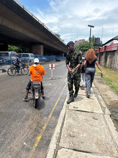 In Ikoyi, Nigeria, under a large overpass, a scene unfolds with several individuals on a paved road. A soldier in camouflage uniform stands near the center, his hands clasped in front of him, looking slightly to the right. To his right, a woman with long, curly reddish-brown hair walks away from the camera, holding a dark object in her right hand. Behind them, another person, a male with short dark hair, walks alongside the woman.
In the foreground, a man on a motorcycle wears an orange hoodie and dark pants, facing away from the viewer. He has a satchel slung over his shoulder. To the left, further down the road, another man cycles a bicycle, also facing away.
The road surface is asphalt, marked with yellow lines, and shows signs of wetness and tire marks. A "NO PARKING" sign is visible on a metal pole. The background features the concrete structure of the overpass, vehicles, and a red-and-white striped fence. Greenery and trees are present beyond the fence, and the sky is partly cloudy. The overall environment suggests an urban setting with traffic and pedestrian activity.