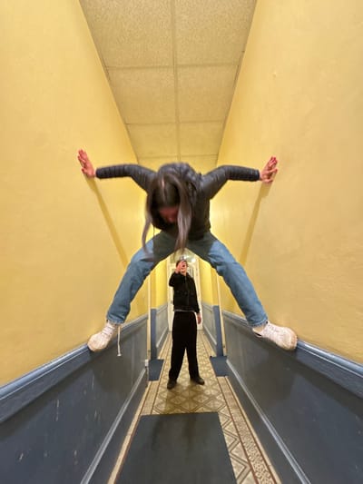 A wide-angle photograph depicts a narrow indoor hallway with two individuals. The hallway features light yellow painted walls with dark blue baseboards. The floor is covered with geometric patterned tiles in beige and dark blue hues, and a dark mat is visible in the foreground. The ceiling consists of a light-colored suspended acoustic tile grid.

In the foreground, a person with long dark hair, wearing a dark puffer jacket, blue jeans, and white athletic shoes with untied laces, is positioned horizontally across the hallway. Their back faces the viewer, arms outstretched and hands pressed against the yellow walls. Their legs are spread, with feet touching the dark blue baseboards on either side, indicating a suspended or braced posture between the walls. Their head is tilted downwards.

Further down the hallway, a second person, dressed in a dark jacket, dark pants, and dark shoes, stands upright. This individual is holding a smartphone up, facing the foreground subject and the camera, actively capturing the scene. The exact location in Roanne, France, cannot be determined from the provided visual information.