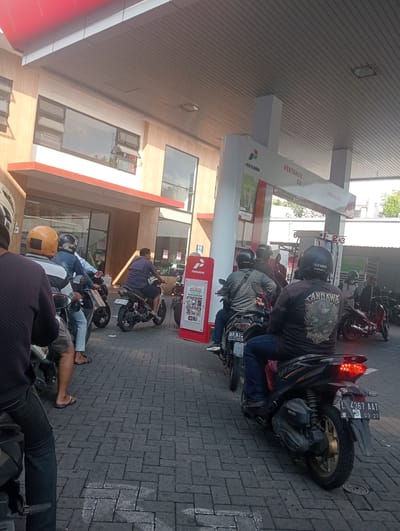 The image depicts a group of people on motorcycles waiting in line at a Pertamina gas station in Surabaya, East Java. The scene is outdoors, under a covered canopy of the gas station. Several individuals are seated on their motorcycles, facing forward, indicating they are waiting for service. The motorcycles are parked in a line on a brick-paved surface. In the background, there is a building with large windows and several Pertamina-branded signs, including "PERTAMAX 92" and "PERTALITE". Some people are wearing helmets, and their attire suggests a casual, everyday setting. The lighting indicates it is daytime, with shadows cast by the canopy and the building. The overall atmosphere suggests a typical queuing situation at a public service location.