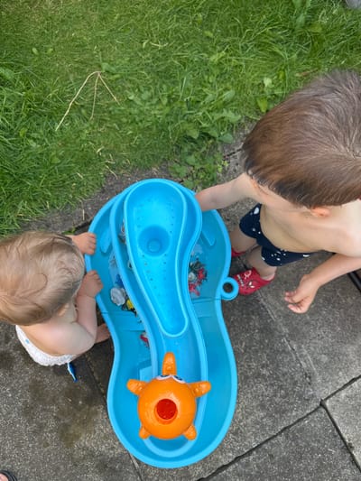 An overhead shot shows two young children playing with a blue water table in an outdoor setting. The water table is filled with water and has various toys, including an orange fish. The ground is a mixture of grass and paving stones.
One child, facing away from the camera, wears a diaper and is seated on the ground, reaching into the water table. The other child, also facing away from the camera, stands and wears dark blue shorts with white star patterns and red Crocs-style shoes. This child's hand is extended towards the water table.
The environment appears to be a backyard, with green grass and grey paving stones. The lighting suggests it is daytime, likely outdoors. There is no specific indication of the city of Leeds, United Kingdom, within the image itself.