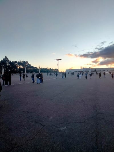 A large open paved area is filled with many people. In the background are trees and various buildings, including a modern rectangular building. A tall, thin crucifix stands prominently in the center of the scene. The sky above is a mix of blue and orange, suggesting either sunrise or sunset, with clouds present on the right side of the image. The light is soft, giving the scene a peaceful and contemplative atmosphere.