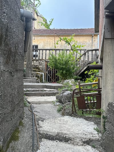 This is a photograph taken in the city of Vranjina, Montenegro. The image shows a narrow, outdoor pathway between two buildings, leading upwards. The pathway is made of concrete steps with rough, gravel-like surfaces. The building on the left is constructed of grey, rough-textured concrete. On the right side of the pathway, there is a rusted metal structure resembling a gate or part of a swing, with wooden slats and a yellow object behind it. Further up the steps, there is a stone wall with a decorative concrete balustrade. A lush green tree grows in front of the balustrade, and beyond that, a building with a yellow facade and a tiled roof is visible. Above the pathway, a grey pipe runs along the concrete wall of the left building, and branches of a vine with green leaves overhang from above. The sky is overcast and light grey. The scene appears to be an outdoor alleyway or courtyard in a residential area, possibly an older part of the city.