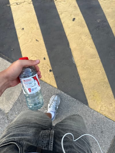 A person's hand holds a Vittel water bottle with a red cap. The bottle has a label that reads "UNE VITALITÉ SOURCE DE POSITIF" and features a graphic of a person running. The person is wearing grey denim jeans and white sneakers. A white cord, possibly from headphones, is visible in the lower right corner. The scene is outdoors on a paved surface marked with alternating black and yellow stripes, characteristic of a pedestrian crossing or caution zone. The image appears to be taken from a first-person perspective. The location is the city of Alès, France.