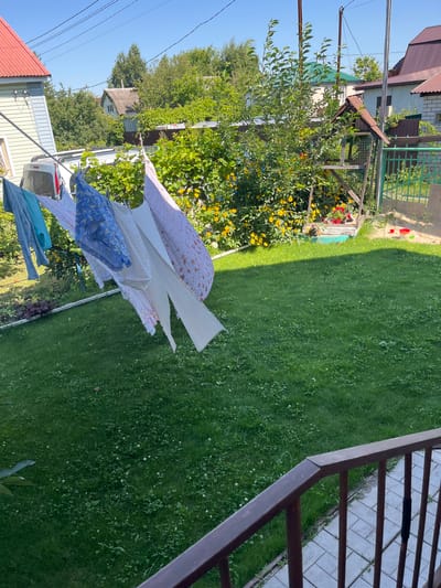 This is a medium shot of a backyard on a sunny day. In the foreground, a brown railing with vertical slats frames the bottom right corner of the image. A paved walkway is visible beyond the railing. The main focus of the scene is a clothesline filled with laundry, stretching across the yard from left to right. Several items of clothing, including a blue t-shirt, a patterned blue and white garment, and a white sheet, are hanging on the line, some billowing in the breeze. Behind the clothesline, a well-maintained green lawn covers the majority of the yard. Further back, there is a dense bush with yellow flowers, and a small wooden structure that appears to be a bird feeder or playhouse. A green metal fence marks the boundary of the property. Several houses and trees are visible in the background, indicating a residential area. The sky is clear and blue, suggesting fair weather. This scene is consistent with a suburban or rural backyard in Kazinka, Russia, likely during the summer months.
