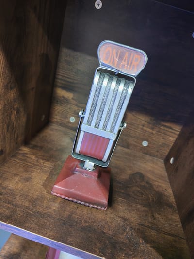 An antique-style microphone, featuring a reddish-brown base, silver mesh grille, and a silver frame, is positioned within a dark brown wooden shelf unit in Picassent, Spain. The microphone's top displays an "ON AIR" sign with orange lettering. The wooden unit exhibits a visible grain pattern and is constructed with multiple silver Phillips-head screws and white circular covers visible on its interior surfaces. No individuals, actions, or direct interactions are depicted.