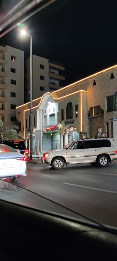 An urban night scene captures a street in Jeddah, Saudi Arabia, from inside a moving or stationary vehicle. The sky is dark, and the surroundings are illuminated by artificial light sources.
In the mid-ground, a distinctive building stands out with a light-colored, stone-like facade, adorned with decorative orange/yellow strip lighting that outlines its roofline and arched architectural features. A prominent sign in red Arabic script, "السيسم" (Al Sism or Al Seasem), is visible above the main entrance, with additional Arabic text "المبتسم" (Al Mubtasim) to its right. Palm trees are visible near this building on the left. Taller, multi-story residential or commercial buildings with visible windows and balconies rise in the background. A street light pole stands prominently in the center-left.
Several vehicles are present on the street. In the foreground on the left, the rear of a white sedan is partially visible. Behind it, a dark SUV is mostly obscured. To the right, a white SUV, possibly a Toyota Land Cruiser, is moving or stopped, with its headlights on. A person can be seen in the driver's seat. The road surface appears damp or reflective under the streetlights.
The overall scene depicts a typical busy night in an urban area, with traffic and illuminated buildings suggesting commercial or residential activity.
