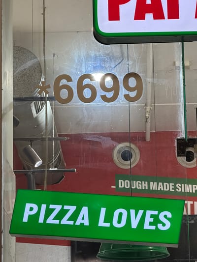 An image captures the exterior view of a storefront, likely a pizza restaurant, in Kfar Saba, Israel. The scene is dominated by illuminated signs and reflections on a glass partition.
On the left, a portion of a vertical red and white sign is visible, displaying the letters "PA". Centrally, a clear glass surface features large, golden-brown numbers "6699" followed by an asterisk "*". Behind this glass, a red wall displays the text "DOUGH MADE SIMPLE" in white letters. Below this text, a circular object, possibly a security camera or sensor, is mounted on the wall. Further left and behind the glass, a white pipe or conduit runs horizontally.
On the right side of the frame, a bright green illuminated sign stands vertically, clearly displaying the words "PIZZA LOVES" in white capital letters. A faint reflection of another sign, partially showing "AT", is visible behind it on the red wall. In the background, to the far right, a dark green cylindrical bin or container can be seen.
Below the main signs and visible through the glass, there is what appears to be an interior space containing a large, cylindrical grey object with vents, possibly an air conditioning unit or a large fan. A small spotlight is also positioned in this area. The glass itself shows some smudges and reflections, indicating it is an outdoor-facing window. No people or specific activities are visible, presenting a static view of a business entrance. The lighting suggests indoor illumination or perhaps evening hours.