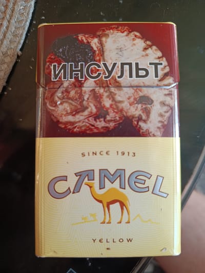 The image displays a close-up, eye-level view of a Camel cigarette pack resting on a dark, textured surface. The pack is predominantly yellow and features the iconic Camel logo with a camel silhouette. The top portion of the pack has a graphic of what appears to be a medical illustration of a brain with text in Cyrillic that reads "ИНСУЛЬТ," which translates to "STROKE" in English. The bottom portion of the pack displays "SINCE 1913" and "YELLOW." A woven object, possibly a straw mat, is visible to the left of the cigarette pack. The background is dark and out of focus. There is no indication of the location being within the city of Прибрежный ауылдық округі, Kazakhstan.