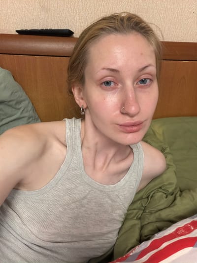 An indoor, close-up shot shows a young woman with fair skin and blonde hair lying on a bed. She is wearing a grey tank top and silver hoop earrings. The background includes a wooden headboard and a textured wall. A dark remote control is visible on top of the headboard. The woman has a nose ring and appears to be looking directly at the camera. Her eyes are blue, and her lips are slightly parted. She is resting on green bedding.