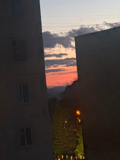 The image depicts a cityscape at dusk, with a dramatic sunset illuminating the sky with vibrant orange and pink hues. The foreground is dominated by the dark, imposing facade of a building, its windows appearing as dark rectangles. Above this structure, a sliver of the sky is visible, revealing a band of colorful clouds stretching across the horizon. Beyond this, a dark, dense mass of trees is illuminated by a warm, orange streetlamp, suggesting an urban or suburban setting. The overall mood is serene and contemplative, capturing the transition from day to night. The scene appears to be set in Neryungri, Russia, though no specific landmarks are identifiable. The image is likely a photograph taken from a slightly elevated position, looking out over the city as the day concludes.