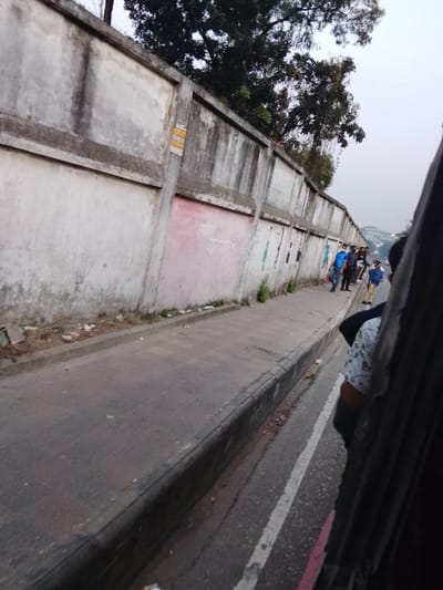 The image captures a street scene in Shibpur, Bangladesh, viewed from a moving vehicle, likely a bus or auto-rickshaw, indicated by the partial view of a passenger and vehicle interior on the right. A long, weathered concrete wall with prominent rectangular panels dominates the left side of the frame, extending into the distance. The wall is discolored with streaks of dark grime and patchy areas of lighter and darker concrete. One section towards the middle features a faded pink paint patch with some illegible writing, possibly in Bengali script. A small yellow sticker or label is affixed to a vertical support on the wall.
Adjacent to the wall is a dark gray paved sidewalk, showing some signs of wear and minor litter at its base. Small patches of green vegetation sprout from the ground along the wall's foundation. A group of people, including men and boys, are visible on the sidewalk further down the street. One man in a blue shirt and a light blue face mask is clearly seen, while others are partially obscured. A young boy in a light blue top and dark pants is also discernible. They appear to be walking or waiting by the wall.
To the right of the sidewalk is the road, marked by a clear white solid line and a fainter red line running parallel to the curb. The sky above is overcast and bright, suggesting daytime, possibly late morning or afternoon. Trees and foliage are visible above and behind the wall, particularly on the upper left and in the distant background. The overall impression is that of an urban or semi-urban setting with everyday activity.