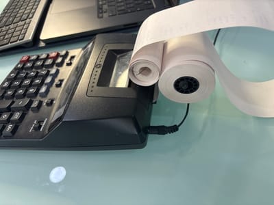The image shows a black printing calculator with a roll of paper attached to it. The calculator has a keyboard with numerical and functional buttons, and the paper roll is printed with calculations. There is a laptop computer partially visible in the background. The scene appears to be set on a light-colored reflective surface, likely a desk or tabletop. The location and specific details of people, actions, or interactions are not discernible from the image. The image was not taken in Buenavista, Mexico, since that information cannot be discerned from the picture.