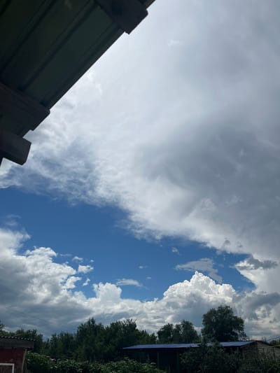 The image captures a cloudy sky over a rural or semi-rural area. In the foreground, on the left side, a dark green corrugated metal roof extends from the top left corner downwards. Below the roof, wooden beams are visible. The sky is predominantly filled with a mix of dark gray storm clouds and brighter white cumulus clouds, with a patch of clear blue sky visible in the center. The horizon line, which is not entirely visible, is obscured by a dense line of green trees and some low-lying vegetation. Further back, there are glimpses of buildings with dark blue and grey roofs, suggesting the presence of structures. The scene appears to be outdoors, possibly in the early stages of a storm, with the sky dominating the upper portion of the frame. There are no people or animals visible in the image. The location is identified as Qobda, Kazakhstan.