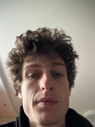 The image presents a close-up, low-angle portrait of a young adult male, likely in his twenties, with fair skin and dark brown, voluminous, curly hair. His gaze is directed slightly downward, and his expression is neutral. His mouth is closed, and his lips are full. Faint lines are visible on his cheeks near his nose and mouth. He is partially clad in a dark, possibly black, garment, the collar of which is visible at the bottom of the frame. The background consists of a plain, light-colored ceiling, with a subtle circular architectural detail or recessed light fixture visible towards the upper right. The scene is an indoor environment. The image itself provides no specific visual cues or landmarks to confirm the location within the city of Vincennes, France, as specified in the request.