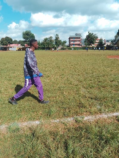 A young man in a camouflage jacket and purple pants walks across a grassy field in Rungiri, Kenya (latitude: -1.249, longitude: 36.692). The field is likely a soccer field, as there is a white line painted across the grass and several figures can be seen in the distance, likely playing soccer. Several buildings, including a red multi-story building with an attached roof, can be seen in the distance. The sky is blue and cloudy, with a few white puffy clouds.