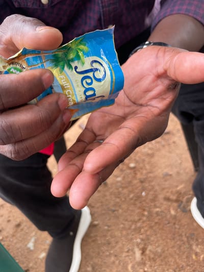 A close-up, eye-level shot captures a pair of dark-skinned hands manipulating a crumpled blue and gold packet. The packet, partially open, displays a palm tree graphic and text that includes "Peal" and "Full Cream". One hand holds the packet open, while the other appears to be reaching into it. The background is blurred but suggests an outdoor setting with reddish-brown ground, possibly dirt or gravel. The person's clothing, visible in the upper portion of the frame, is a dark, patterned shirt. The lower portion shows dark pants and a white-soled shoe. The scene is consistent with an individual in Jimeta, Nigeria, consuming a packaged food product.