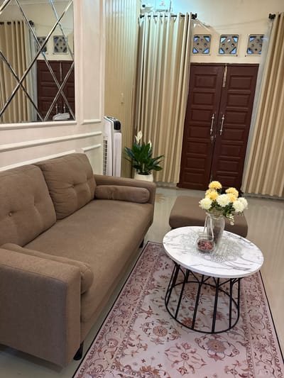 This is a medium-shot interior photograph of a living room, likely located in a home in the city of Pematang Siantar, Indonesia. The room is furnished with a modern, earth-toned sofa on the left, facing a round marble-top coffee table with a black metal base. On the coffee table, a glass vase holds a bouquet of artificial yellow and white flowers, and a small glass jar contains dried red potpourri. Next to the coffee table, to the right, is a small, square brown ottoman.
Dominating the background is a pair of dark brown double doors, possibly leading outside or to another part of the house, flanked by cream-colored curtains. Above the doors, three square decorative vents are visible. To the left of the doors, a tall, white tower fan stands near a large mirror with a diamond-shaped paneled frame, reflecting the room and a portion of the doors. A white plant in a pot is situated to the left of the ottoman, between the sofa and the doors. The floor is covered with a light-colored rug featuring a repeating floral pattern in shades of pink, red, and brown. The walls are painted a light neutral color, with white wainscoting on the lower portion near the sofa and mirror. The lighting appears to be a mix of overhead and natural light, creating a well-lit but slightly warm ambiance. No people are visible in the image.