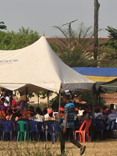 The image captures an outdoor gathering in San Martín, Colombia, under a large white, peaked tent. In the foreground, a young adult male, wearing a blue and black striped short-sleeved shirt, dark trousers, and dark shoes, walks from left to right on a patch of dry, sparse grass. Under the tent, a crowd of individuals is seated on blue, green, and orange plastic chairs, primarily facing an unseen central point. Many are dressed in vibrant, patterned clothing and head coverings; several men are observed wearing red or orange hats. The white tent bears partially legible black text, including "NATION BY RBH" and "NO HOMES." To the right of the main tent, a section of a blue and yellow fabric structure is visible. The background features dense green trees and shrubs, including a tall palm tree on the right, and the reddish-brown roof of a distant building. A solar-powered street light fixture is visible above the foliage. The sky is uniformly light gray.