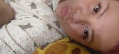 The image is a close-up, rotated, indoor photograph of a young person lying down. The person is looking towards the camera, with their face angled upwards. Their eyes are open, and they are wearing makeup, including eyeliner and mascara. Their lips are slightly parted, and they have a small stud earring in their visible ear. The person is wearing a white garment with a lace-like pattern and what appear to be small gray animal figures printed on it. They are resting their head on a textured surface that is yellow and brown, possibly a blanket or pillow. The background is out of focus but appears to be a domestic setting. There are no indicators or landmarks visible in the image that would place it in Marynivka, Ukraine, or any specific city.