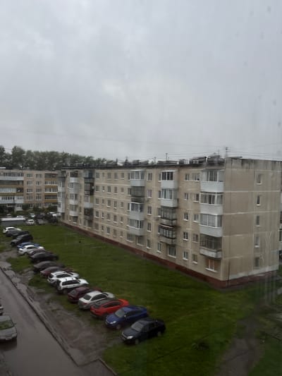 The image shows a cloudy, overcast day in a residential area in Kamensk-Uralsky, Russia. The scene is dominated by two tall, multi-story apartment buildings with beige and grey facades. The buildings have numerous windows and balconies, many of which are enclosed. In front of the buildings, a grassy area is visible, with a row of parked cars extending along the side of the street. The street itself appears wet, suggesting recent rain. The cars are of various colors and models, and are parked in a somewhat orderly fashion. The sky is a uniform grey, and trees with green foliage line the top of the apartment buildings. There are electrical wires visible in the sky.