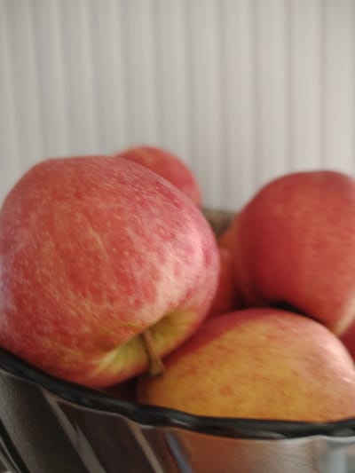 The image shows a close-up view of a bowl filled with red and yellow apples. The apples are the primary subjects, with their textured skins and rounded forms filling most of the frame. The bowl appears to be made of dark, possibly metallic material with a reflective surface. In the background, there is a blurred white surface with vertical lines, suggesting a wall or a blind. There are no people or any signs of human activity. The lighting is soft, casting gentle shadows. The scene is indoors and appears to be a still life. The location is noted as Кривец, Russia.