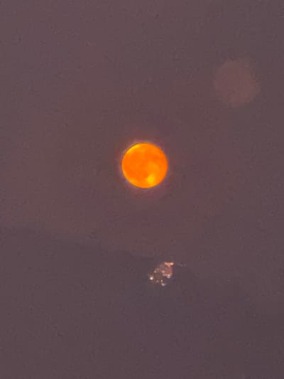 An orange, luminous sphere, possibly the sun obscured by atmospheric conditions, is centrally located in the frame against a dusky, purplish sky. Below and slightly to the right of the sphere, a cluster of small, bright lights, indicative of a distant cityscape or industrial area, is visible. The image is a photograph taken in Balerna, Switzerland.