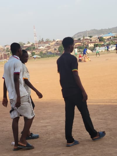 Three young men are standing in a dirt field in Ibo, Nigeria. One is wearing a black shirt and pants with blue sandals. Two others are wearing white shirts, light-colored shorts, and sandals. In the background are buildings and a communication tower. There are other people in the background engaged in what appears to be soccer.