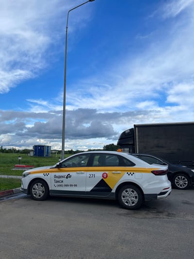 The image shows a white Yandex Go taxi with yellow stripes parked on an asphalt surface in what appears to be Бавыкино, Russia. The taxi has the number 271 on the side and the phone number (495) 999-99-99. Behind the taxi, a dark-colored truck is partially visible. The sky is blue with scattered white clouds. A street light pole stands to the left of the taxi. The environment includes green vegetation.