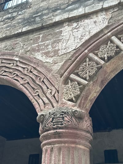 The image shows a close-up of ornate stone carvings on an architectural element, likely part of a building's facade or interior. The stone is a reddish-pink hue, showing signs of age and wear with some chipped areas and exposed underlying brick. The carvings are intricate and detailed, featuring geometric patterns and stylized motifs. On the right side, there are several circular designs with radiating lines and smaller circular embellishments, resembling stars or elaborate crosses. To the left, a series of wavy, interlocking lines form a decorative band. The top of the image reveals a section of aged brick wall with a stone lintel above, and a glimpse of a barred window. The lower portion of the image shows a portion of a fluted column with more sculpted detail at its capital, topped by volutes and a band of complex, layered carvings. The background is a dark recess, suggesting an archway or covered space. This architectural detail is located in the city of Huejotzingo, Mexico, specifically at the Monastery of San Miguel Arcángel, a historic Augustinian convent.