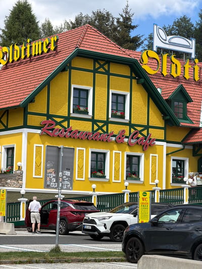 This is a vertical, eye-level shot of a bright yellow, traditional Austrian restaurant and cafe, "Adtimer," with a red tile roof and dark green trim. The facade features several windows with dark green shutters and red flowers in window boxes. A large, stylized yellow sign with dark green outlines spells out "Adtimer" along the roofline. A red script sign spelling "Restaurant & Cafe" is mounted vertically on the yellow facade.
In the foreground, parked cars line the street in front of the restaurant. A dark gray SUV and a red compact SUV are visible. A man in a white polo shirt and light-colored shorts is standing next to the open driver's door of the red SUV, appearing to be exiting or entering. A yellow sign with German text, including "WC GRATIS FREE LIBRE," is visible near the parking area.
The scene suggests a sunny day, with a clear blue sky and some fluffy white clouds visible through trees. The overall impression is of a charming, well-maintained establishment in a picturesque setting, likely a tourist destination given the multilingual "free WC" sign. The architecture and signage point to a location in Austria, as indicated by the provided context.