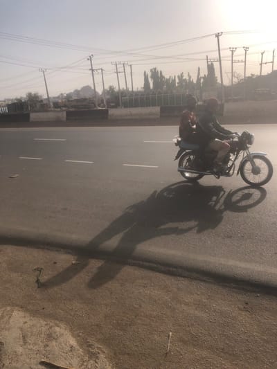 Two adult males are observed riding a silver and dark-colored motorcycle on a paved two-lane road in Hanwa, Nigeria. The driver wears a dark jacket, light trousers, and a red helmet. A passenger, dressed in a dark red shirt, sits behind the driver. Both individuals and the motorcycle cast prominent, elongated shadows across the dark asphalt road surface and onto the lighter brown dirt shoulder, indicating a low sun angle. The road features three dashed white lane markers. To the left of the road, a low concrete barrier with a white top and dark base separates the roadway from a vegetated area containing multiple wooden utility poles with overhead power lines, various trees, and a distant rocky hill, all partially obscured by atmospheric haze.
