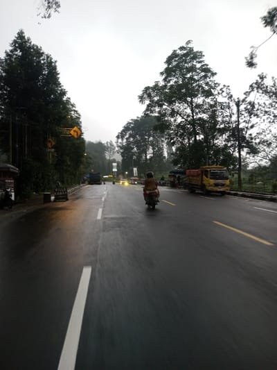 The image depicts a wet, two-lane asphalt road under an overcast, light gray sky, indicating recent precipitation. The scene is located in Dawathang_Dorjibi_ Kashingtsawa, Bhutan.

**Road and Markings:** The road, viewed from a low perspective, is visibly wet, reflecting light. A solid white line defines the left edge of the visible lane, and a broken white line acts as the lane divider. Further down the road on the right, double solid yellow lines and a single solid yellow line are visible, marking additional lane boundaries.

**Vehicles and People:** A lone individual on a dark-colored motorcycle, wearing light brown or tan outerwear, is traveling away from the viewer in the right lane. On the right shoulder, a yellow utility truck with a red open bed is parked or moving slowly. On the left shoulder, a dark blue or black truck is parked. In the distance, several vehicles are visible with illuminated headlights.

**Left Roadside:** The left side of the road features dense green vegetation, primarily tall trees. A utility pole carries wires, and two yellow diamond-shaped traffic signs with black arrows indicating a right curve are mounted on it. A dark-roofed structure, possibly a stall, is present. A white sign with "VILLA KAMARI" partially visible stands on the curb. A dark scooter or motorcycle is parked nearby.

**Right Roadside:** The right side of the road is also lined with trees and vegetation, separated from the road by a low concrete barrier or wall. Beyond the yellow truck, faint figures and indistinct roadside structures are discernible. A white rectangular sign on a pole is visible further down the road.

**Environment:** The sky is uniformly overcast and light gray. The distant horizon is obscured by atmospheric haze. The area is heavily vegetated with numerous tall, mature trees.