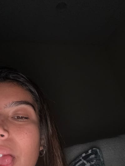 The image presents a close-up, partial view of a female subject in a low-light indoor setting. The lower-left quadrant of the subject's face is visible, featuring medium-brown skin. Her right eye is partially closed or directed downwards, revealing a dark brown iris and a distinct eyebrow. The bridge of her nose is partially visible. Her mouth is open with her tongue extended slightly from the lower-left edge of the frame. Long, dark brown hair frames the right side of her face, and a small, silver-colored hoop earring is present in her right earlobe. The upper and right portions of the image are predominantly dark. In the bottom-right corner, a fragment of a patterned fabric, consistent with a cushion or blanket featuring a light design on a dark background, is visible. There are no other individuals or direct interactions discernible. The environment is dim, with light primarily illuminating the subject's face. The specific location within the city of Princeton, United States, cannot be determined from the visual information provided in this image.