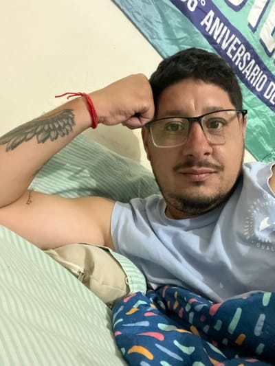 An adult male is depicted in a close-up, chest-up view, appearing to be lying down in an indoor setting. He has dark hair, a short beard and mustache, and wears black-framed eyeglasses. His right elbow is bent, with his right hand supporting the side of his head. A red string bracelet is visible on his right wrist, and a large wing-like tattoo is present on his right bicep. A smaller text-based tattoo is visible on his left bicep. He is wearing a light blue sleeveless t-shirt with a partially visible white graphic on the right chest that includes "BREEZE SURF CO" and "SIXTY". He is resting on light green and white striped bedding, with a beige pillow visible beneath his left arm. A dark blue blanket with a pattern of colorful abstract shapes covers his lower torso. In the background, a light-colored wall is visible, with a teal banner or flag displaying partial white text, including "6° ANIVERSARIO DE," suggesting a celebratory context. The scene is located in Bernal Este, Argentina.